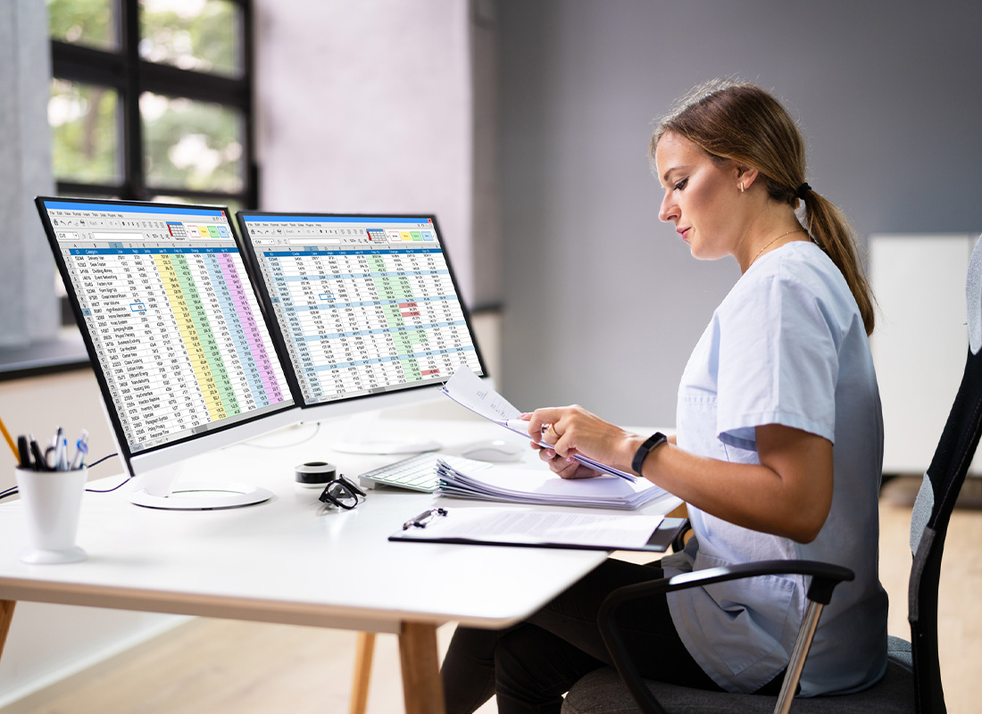 A medical billing professional sits at a computer with two monitors and reads paperwork, illustrating a healthcare reimbursement concept.