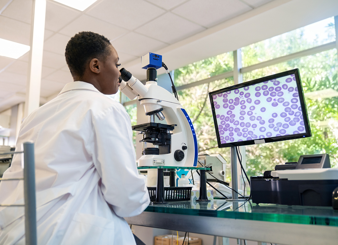 Rear view of a pathologist looking at a sample through microscope with magnified image seen on computer screen, illustrating the concept of AI in pathology.