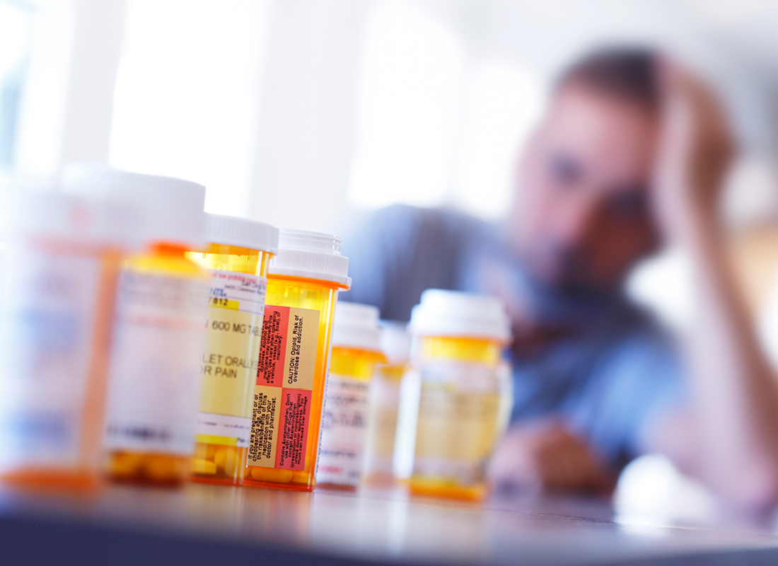 A large group of prescription medication bottles sit on a table in front of a distraught man who is leaning on his hand as he sits at his dining room table. The image is photographed with a very shallow depth of field with the focus being on the pill bottles in the foreground, illustrating the concept of opioid abuse.