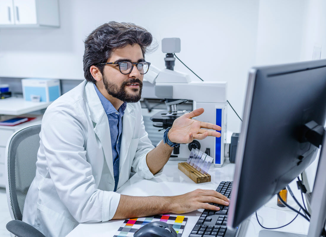 A pathologist wearing a lab coat and glasses works at a computer screen next to a digitally enabled microscope and a tray of glass slides.