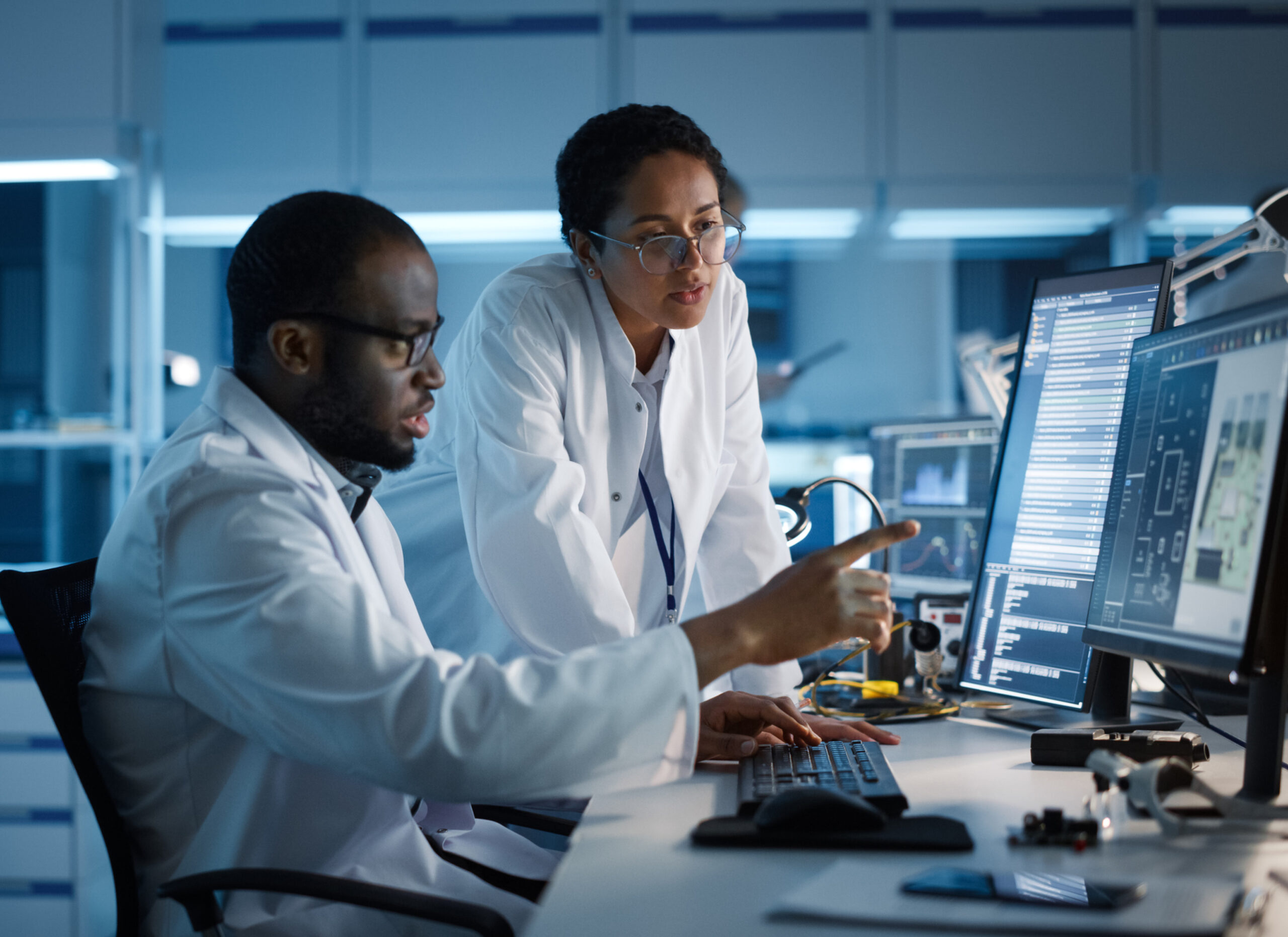 Two laboratory professionals, one seated and one standing, look seriously at computer monitors at a desk in a darkened laboratory, illustrating a cybersecurity concept.