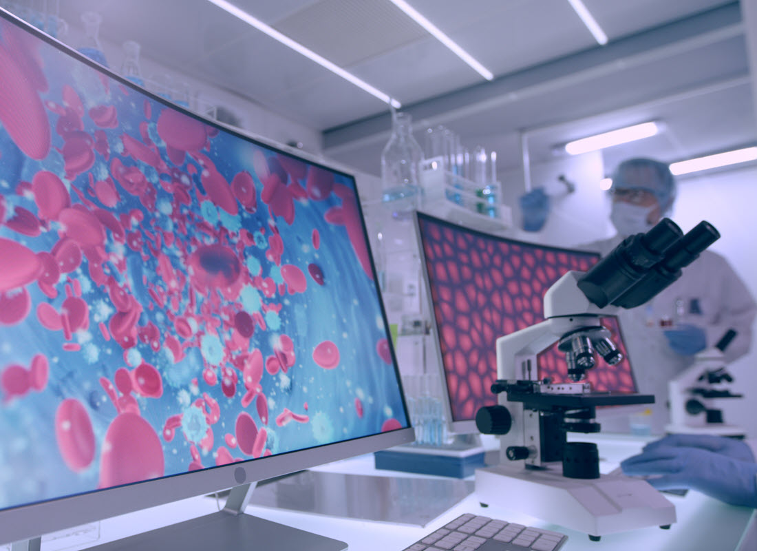 A close-up photograph of an optical microscope between two monitors displaying pathology-related imagery such as red blood cells.