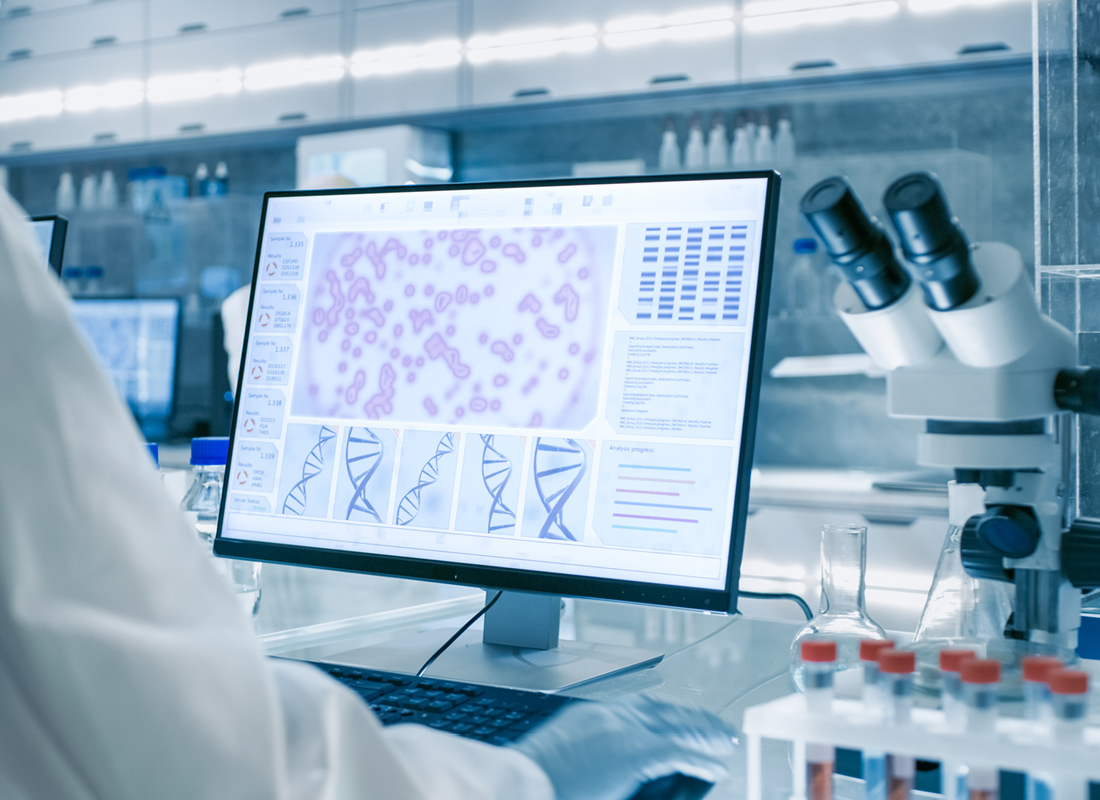 A laboratory technician in a white lab coat works at a computer displaying digital images of cells, gels, and DNA structural models.