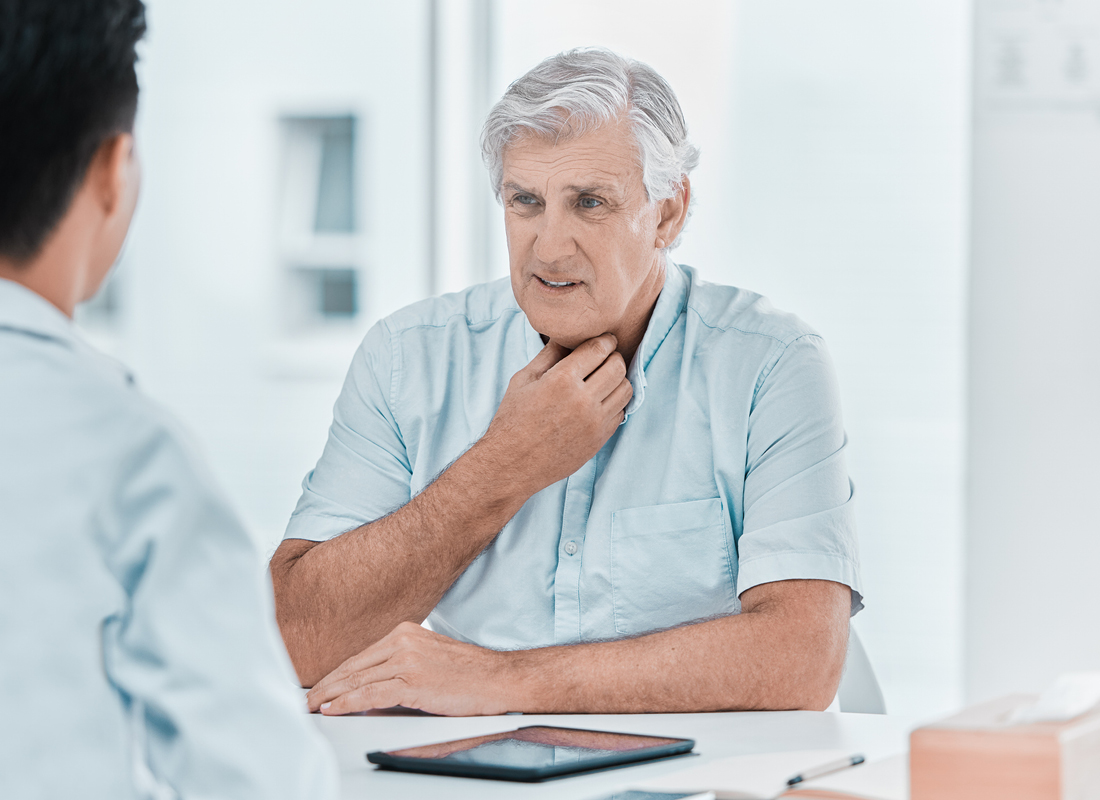 An elderly male patient holds a hand to his throat as he speaks with his doctor, illustrating the concept of throat problems and potential esophageal cancer.