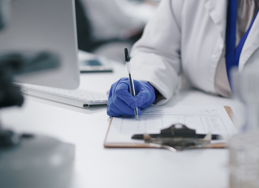 Closeup of a laboratory professional in PPE sitting at a desk with a computer monitor and filling out a form on a clipboard.