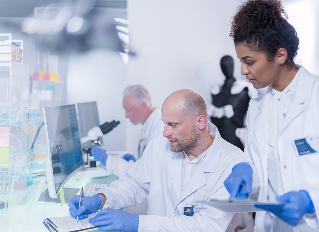 A female lab professional holding a clipboard stands behind her male colleague, who is seated at a lab bench with a computer monitor and paper notebook, illustrating the concept of reviewing laboratory health and safety plans.