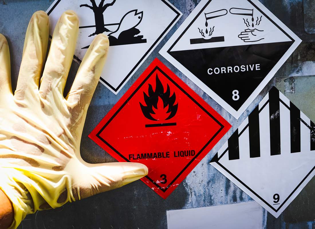 A hand wearing a latex glove rests on the metal side of a container with several warning labels indicating that the contents are flammable, corrosive, and hazardous to the environment.