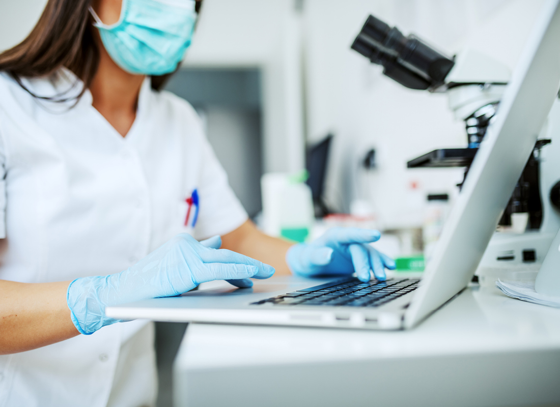 A closeup image of a female laboratory assistant in PPE typing on a laptop at the laboratory bench