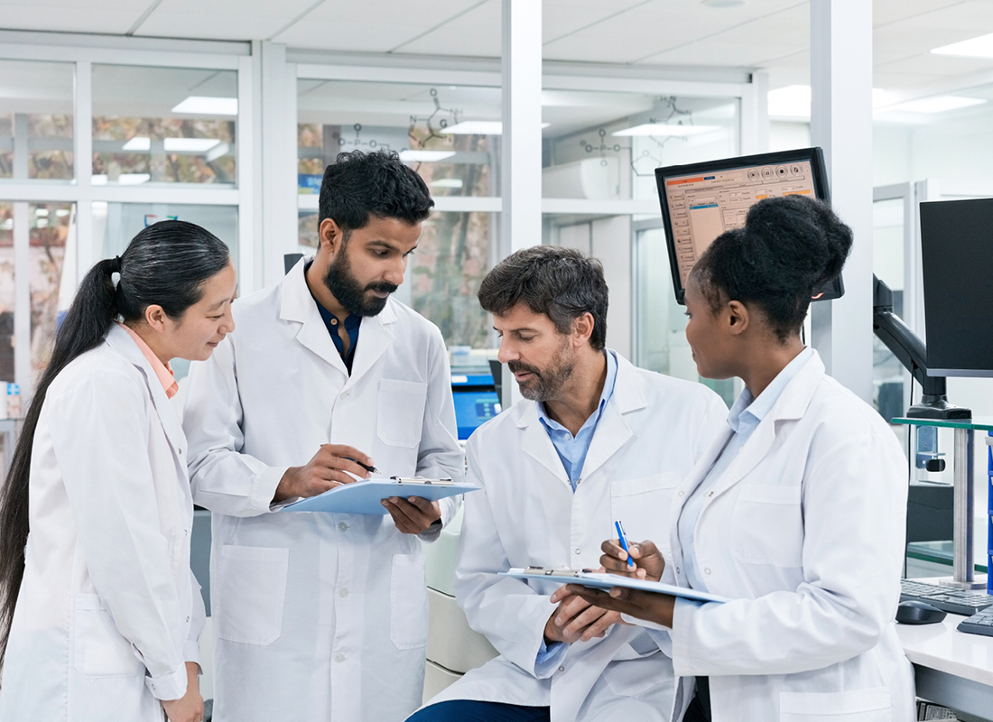 A group of four laboratory professionals, three standing and one sitting, meet in the laboratory. Two of the lab professionals are holding clipboards.