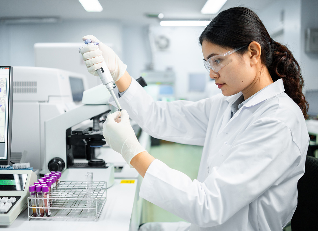 A female laboratory professional pipettes at a lab bench with a microscope, rack of test tubes, and monitor displaying digital pathology images nearby.