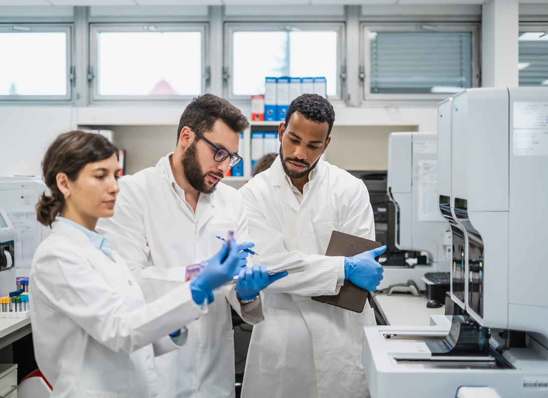 A group of lab technicians, with a mix of genders and ethnic backgrounds, stand focused as they use a digital tablet to review data while engaging with sample analysis equipment in a bright laboratory setting.