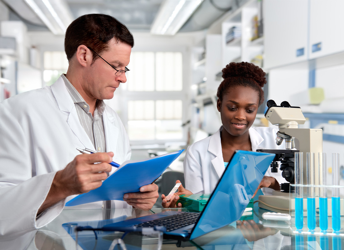 Two laboratorians in white lab coats work together at a bench. One sits at a microscope with a box of glass slides, holding one in her hand. The other is supervising, with a laptop open in front of him; he has a document folder in one hand and a pen in the other.