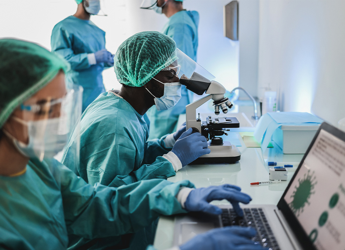 An image of several clinical laboratory scientists in lab coats, hairnets, masks, face shields, and nitrile gloves. A laboratorian in the foreground is working on a laptop; behind her is another using a microscope; two more are partially visible in the background.