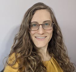 A headshot of Julianna Ianni, a fair-skinned woman with dark blonde hair wearing a mustard yellow shirt and rectangular tortoiseshell glasses.